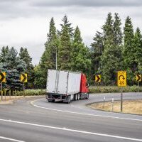 Industrial bonnet bright red classic big rig semi truck transporting commercial cargo in dry van semi trailer turning on the round highway exit with green trees on the side and road signs